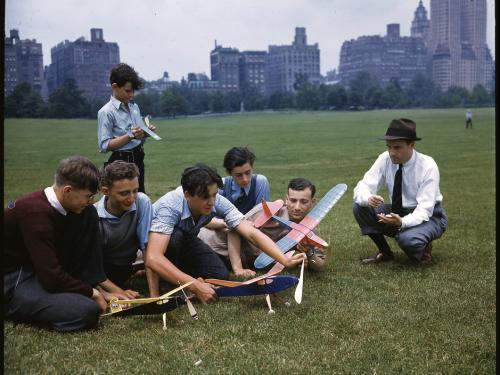 A group of six young men of various ages gather around three model airplanes in a park with a city skyline in the distance. To the right is an adult crouching on the ground next to them with what seems to be either a stopwatch or notebook. 