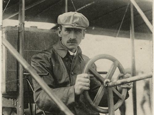 A man sits behind the steering wheel of a biplane, looking toward the camera. The photo looks old and is dated August 1909.