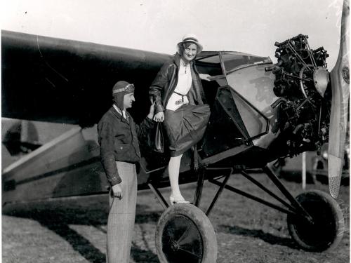 A woman looks toward the camera as she climbs through the door of an aircraft. A man to the left, Clyde Cessna, assists her.