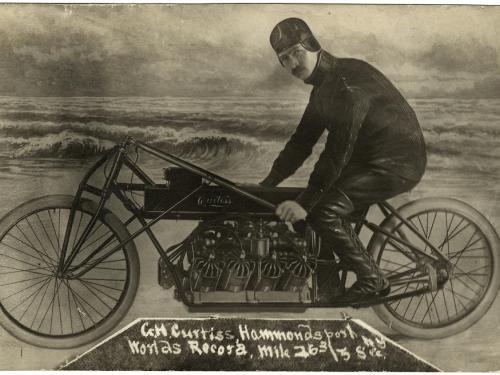 A man sits posed on a motorcycle, looking at the camera. He is in front of what is presumably a fake background meant to look like the beach.