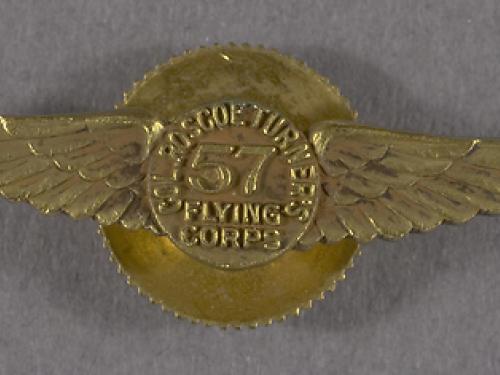 A gold pin in the shape of wings with the number 57 at the center. Around the 57 it reads Col. Roscoe Turner's Flying Corps.