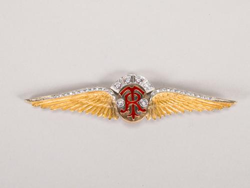 A gold and silver pin in the shape of wings with the letters T and R on top of each other in the center in red. The tops of the wings are encrusted with gems. 