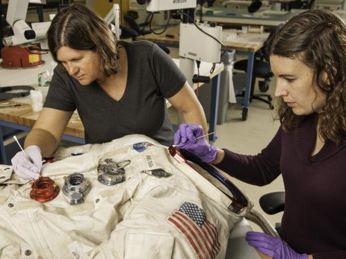 Two women next to a spacesuit working to conserve it with tools.