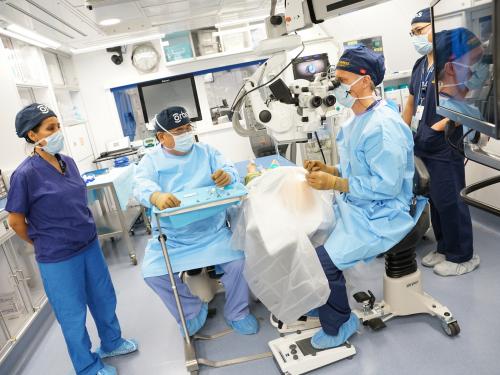 Four medical professionals stand around an operating table. One of them sits next to the table looking into a machine that is focuses on the patient.