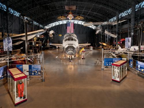 At the center of the photograph is the nose of the Space Shuttle, with the Space Shuttle expanding behind it. Hanging overhead is a satellite, a spacesuit, and a missile. Surrounding on the ground are cases with space stuff. 