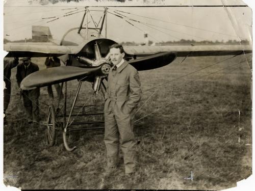 A man in a flying suit stands in front of an aircraft in a field. 