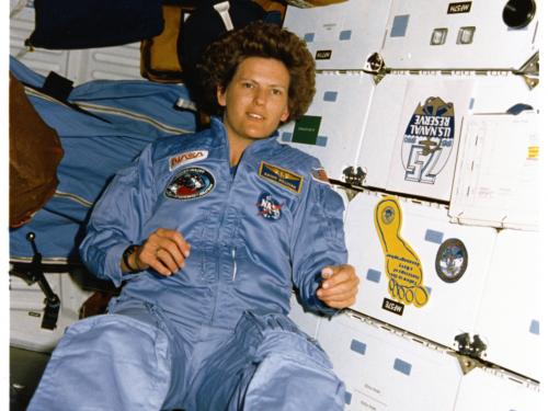 Woman in a light blue NASA flight suit floats weightlessly in front of the white lockered walls of the Space Shuttle.