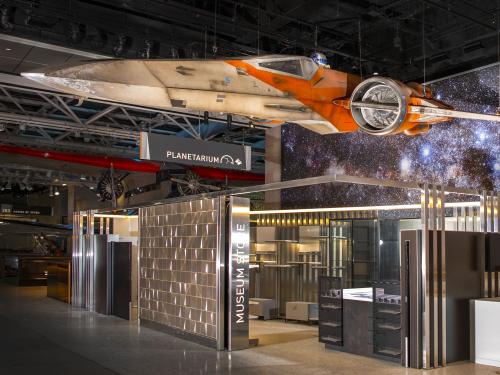 X-wing, a weathered cream and orange space vehicle hangs over an unstocked store and ticket area.