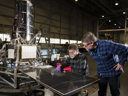 Two men look at a shiny plate on a piece of spacecraft. 