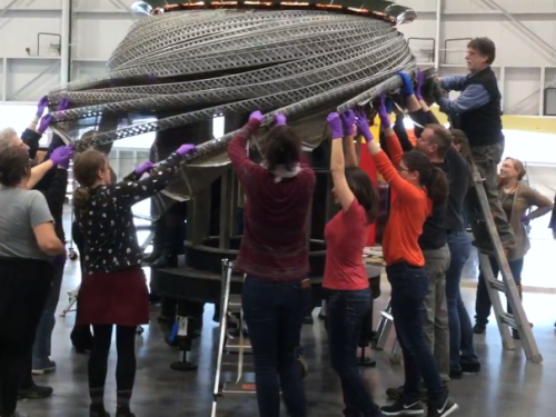 A group of approximately 10 individuals lifting a large object above their heads in a open interior space.