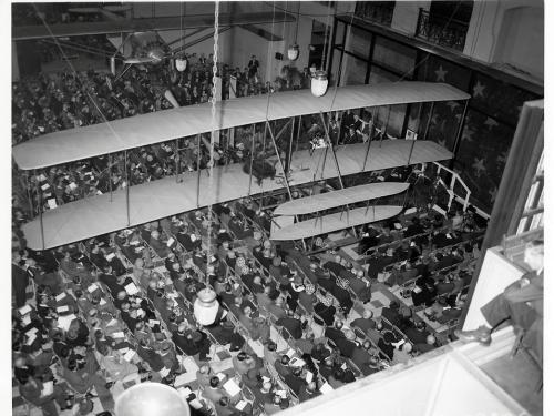 A photo taken from a balcony looking down at a crowd of seated people. The Wright Flyer hangs overs head, with the Spirit of St. Louis hung across from it.