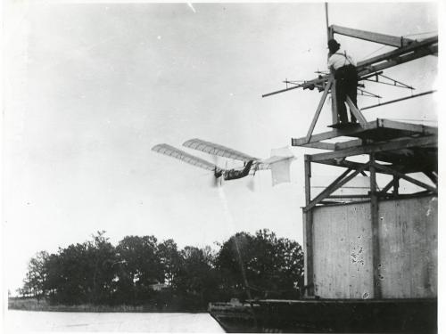 One-half left rear view of a Langley Aerodrome No. 5 in flight over the Potomac River just after launch. A man watches from a houseboat at top right.