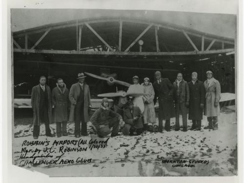 A group of eleven people stand in a line next to each other in front of an aircraft hangar. An airplane can be seen directly behind the group.