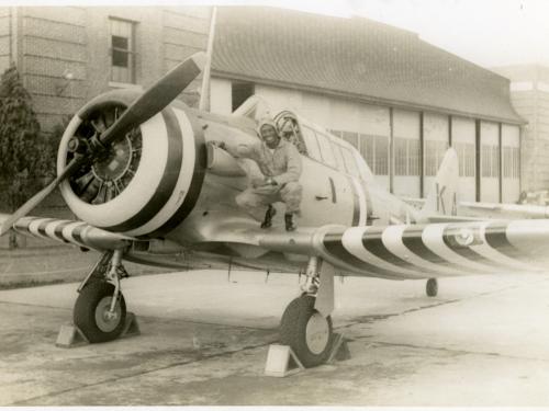 Sepia toned image of Jesse Leroy Brown squatting on the right side wing of an aircraft as he poses for a picture.