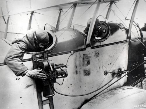 A man reaches out from the back seat of a biplane to look into the viewfinder of a camera that is strapped to the side of the aircraft.