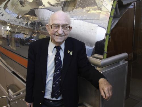 Balding elderly man wearing glasses stands beside a silver reflective airplane nose.