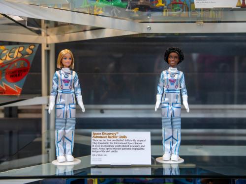 Two Space Discovery Barbies photographed in a glass case. They appear to be standing up straight. Between them is a small artifact description card.