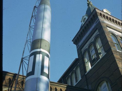 The top three-quarters of green and white missile seen from the ground looking up. The rocket sits vertically in front of an ornate building.