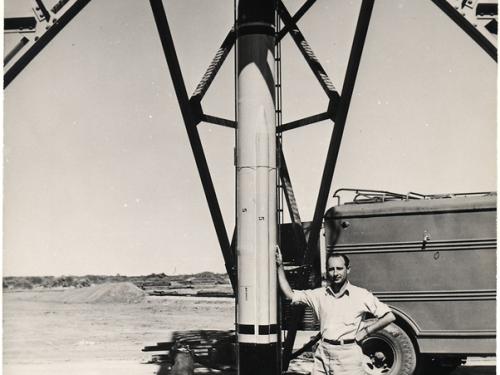 A man leans on a rocket that is about two times as tall as him. The rocket is fixed to a launch tower. Part of a truck can be seen in the background.