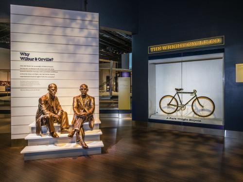 A photo looking into the entrance of a museum exhibit. A bicycle can be seen on display behind plexiglass and a statue of two men sitting adorn the entryway panel which says "who were wilbur & orville."