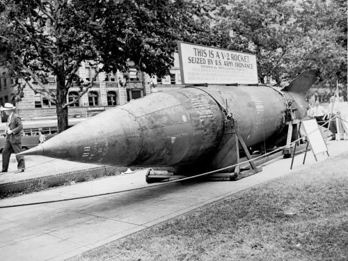 A large rocket lays horizontally on a city street with ropes around it. A sign on top of it states that it is a captured German V-2 rocket.