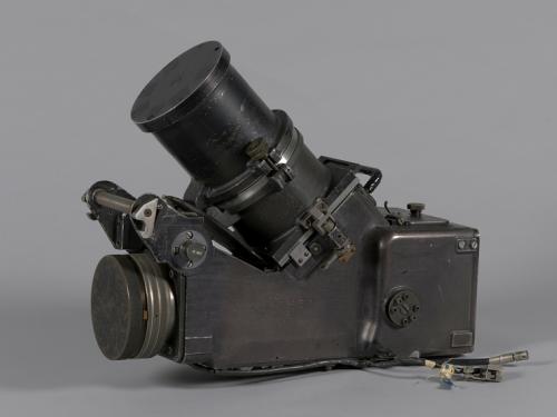 A black object similar in shape to a camcorder. It has a rectangular body with a lens on one end. Its also has another lens sticking out of the top.