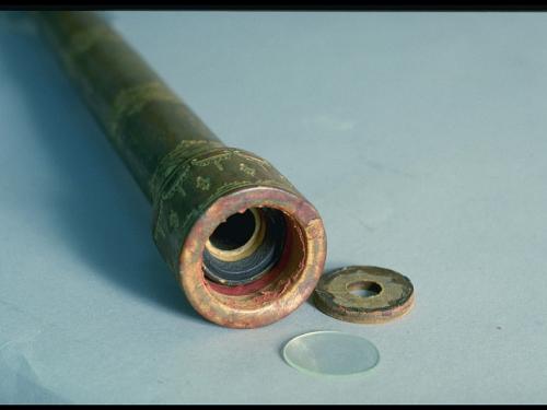 A photo of the end of a simple wooden telescope embellished with golden filigree painted on its surface. The lens and end cap are laid on the table next to it.