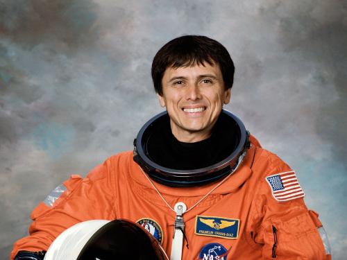 An astronaut holding a helmet in his hand and wearing a bright colored spacesuit smiles for a portrait.