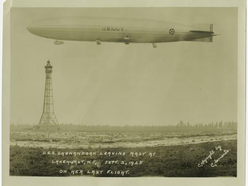 An airship floats in the air at the top of the frame. A mooring mast can be seen in the distance behind it.