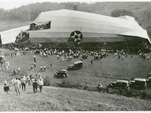 A wrecked piece of an airship sits in a fields as swarms of people flock to it.