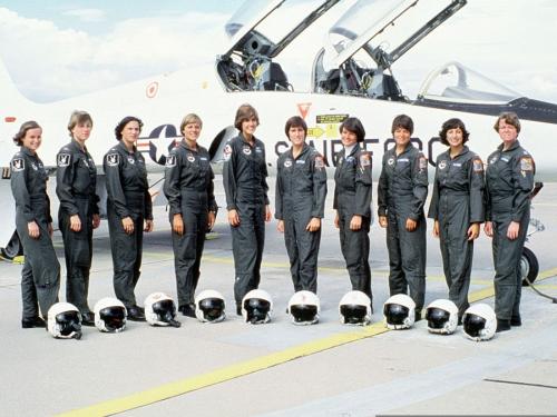 A group of 10 women wearing flight suits pose in front of a fighter jet.