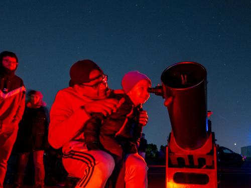 An child looks through a telescope with a thrilled expression while sitting on an adult's lap.