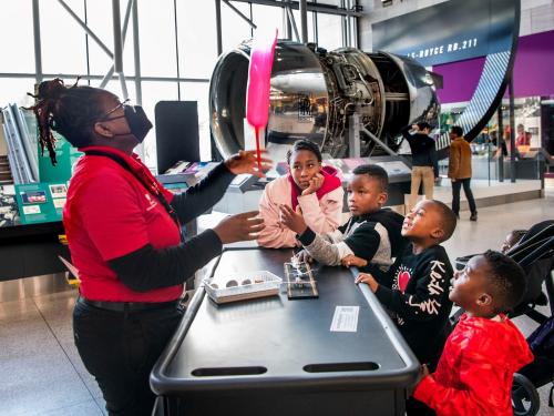 A group of children gather around a mobile desk while an individual does a demonstration using various items.