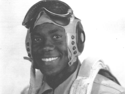 A man poses for a portrait wearing a flight cap and goggles and a flight suit.