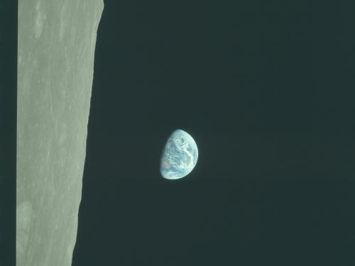 A view of Earth from space, with a large portion of the moon’s surface in the foreground. The Earth appears partially illuminated by the sun, displaying swirls of white clouds over blue oceans and brown land masses.