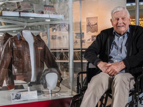 An elderly individual in a wheelchair smiles while seated next to a display of aviation memorabilia, including a leather jacket and a pilot helmet, inside a museum setting.