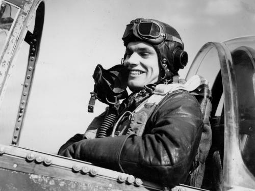 A joyful pilot in a leather jacket and helmet sitting in the cockpit of a vintage aircraft, holding a microphone near their face.