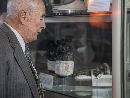 An elderly individual, Bill Anders, looks at a watch on display in a case amongst other space artifacts including gloves and helmets.