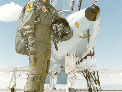 A woman in her mid-20s, wearing a pilot jumpsuit, stands in front of a military aircraft.