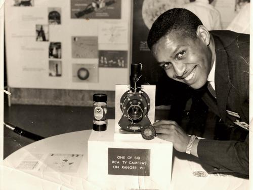 A person smiling at a booth with a display labeled "ONE OF SIX RCA TV CAMERAS ON RANGER VII", showcasing a camera and other equipment.