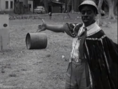 Old photograph on a man pointing toward a small barrel on the ground in a field.