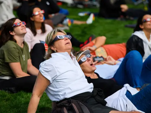 Two people recline on lush grass. They wear eclipse glasses and stare in wonder upward. One person's mouth is open in awe. Behind them are other people in eclipse glasses. 