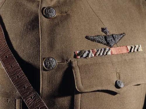 A single breasted drab wool coat with five brass buttons down front; two upper patch pockets and two lower patch pockets with brass buttoned flap; a stiff or "choker" collar; a single gold officer's lace on each cuff; a brass "U.S." insignia on right collar; a Winged Propeller Air Service insignia on left collar; three overseas service chevrons on left cuff; a Distinguished Service Cross ribbon with 7 oak leaf clusters, a French Legion of Honor ribbon, and a French Croix de Guerre ribbon with 3 palm leaves.