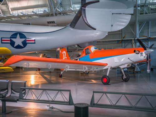 Side view of agricultural plane painted like an orange and white anthropomorphic animated plane, with eyeballs in the cockpit. It sits below a large grey aircraft