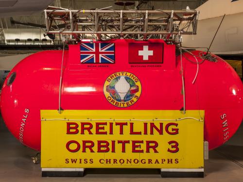 Bright red and yellow metal structure that is the gondola used to carry people and equipment during a balloon ride. It has the inscription, Breitling Orbiter 3. Attached to the metal structure are silver tanks.