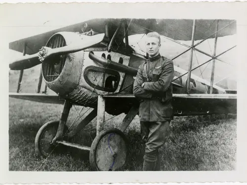 A young white man in a World War I military uniform poses with his arms crossed in front of a biplane.