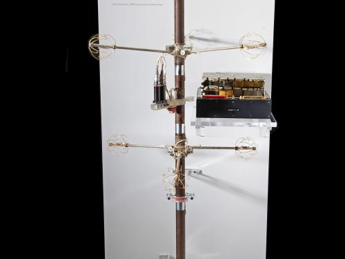 Full view of a long brown stick with three interconnected circles at the top. Two sticks extend horizontally out of the main stick with golden rings at their ends. Silver machinery is at the bottom of the stick.