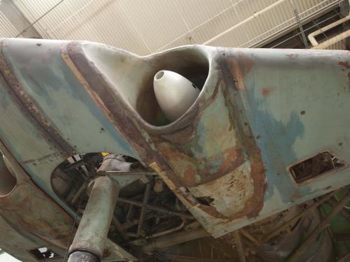 A deteriorated bottom section of a World War II era aircraft shows issues with paint loss, paint discoloration, thinning of metal, and coatings of preservatives visible across the aircraft.