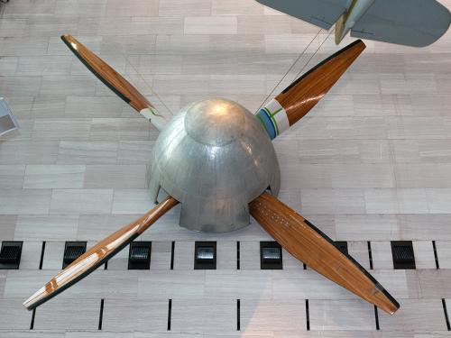 Large, four-blade wooden fan used for a wind tunnel that is hanging in the museum. The center is a silver-colored metal base.