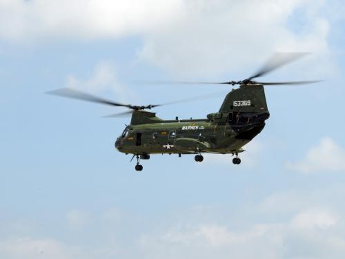 Side view of green military helicopter in flight. The helicopter has two separate propellors, both of which have three blades.